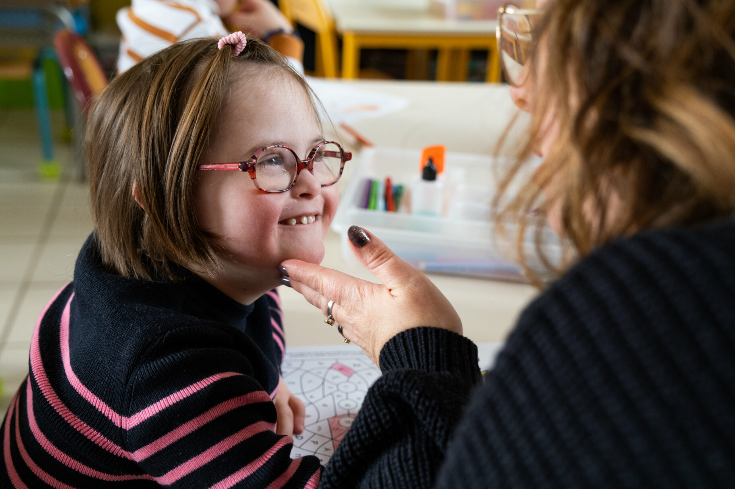 Une petite fille trisomique regarde tendrement son auxiliaire de vie scolaire qui lui caresse le visage. Photographie par Audrey GUYON, photographe du handicap en Normandie.